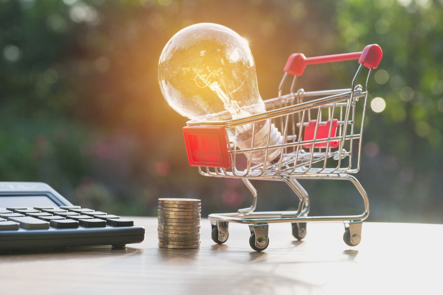 A shopping cart with lighted light bulb beside stack of coins and calculator